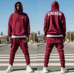 A man wearing a maroon hooded sweatsuit with a small white 'X' logo on the chest of the hoodie front and a prominent white graphic text 'STACKED!' across the shoulders on the back.