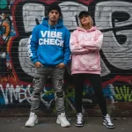 Two models posing against a graffiti wall wearing oversized streetwear hoodies. The man wears a bright blue pullover with large, 3D puff print "VIBE CHECK" text, while the woman wears a light pink hoodie with a detailed, embossed puff print dragon design.