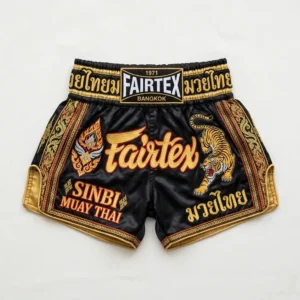 Front view of black Fairtex Muay Thai shorts with a large gold embroidered logo, red accents, and a traditional gathered waistband.