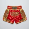 Front view of crimson red and gold Legend Fightwear Muay Thai shorts featuring large embroidered typography and traditional Thai lettering.