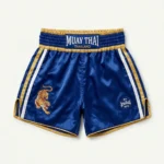 Front view of royal blue satin Muay Thai shorts with a traditional gold waistband patch, side tiger graphic, and gold lettering.