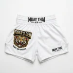 Front view of white Tiger's Den Academy Muay Thai shorts with an orange tiger logo, black waistband patch, and side split hems.