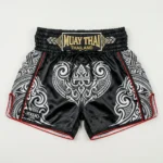 Front view of Yokkao black Muay Thai shorts featuring a white tribal pattern, red piping, and a gold embroidered waistband patch.
