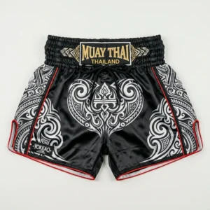Front view of Yokkao black Muay Thai shorts featuring a white tribal pattern, red piping, and a gold embroidered waistband patch.