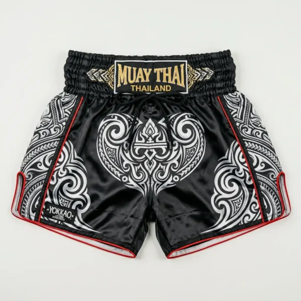 Front view of Yokkao black Muay Thai shorts featuring a white tribal pattern, red piping, and a gold embroidered waistband patch.